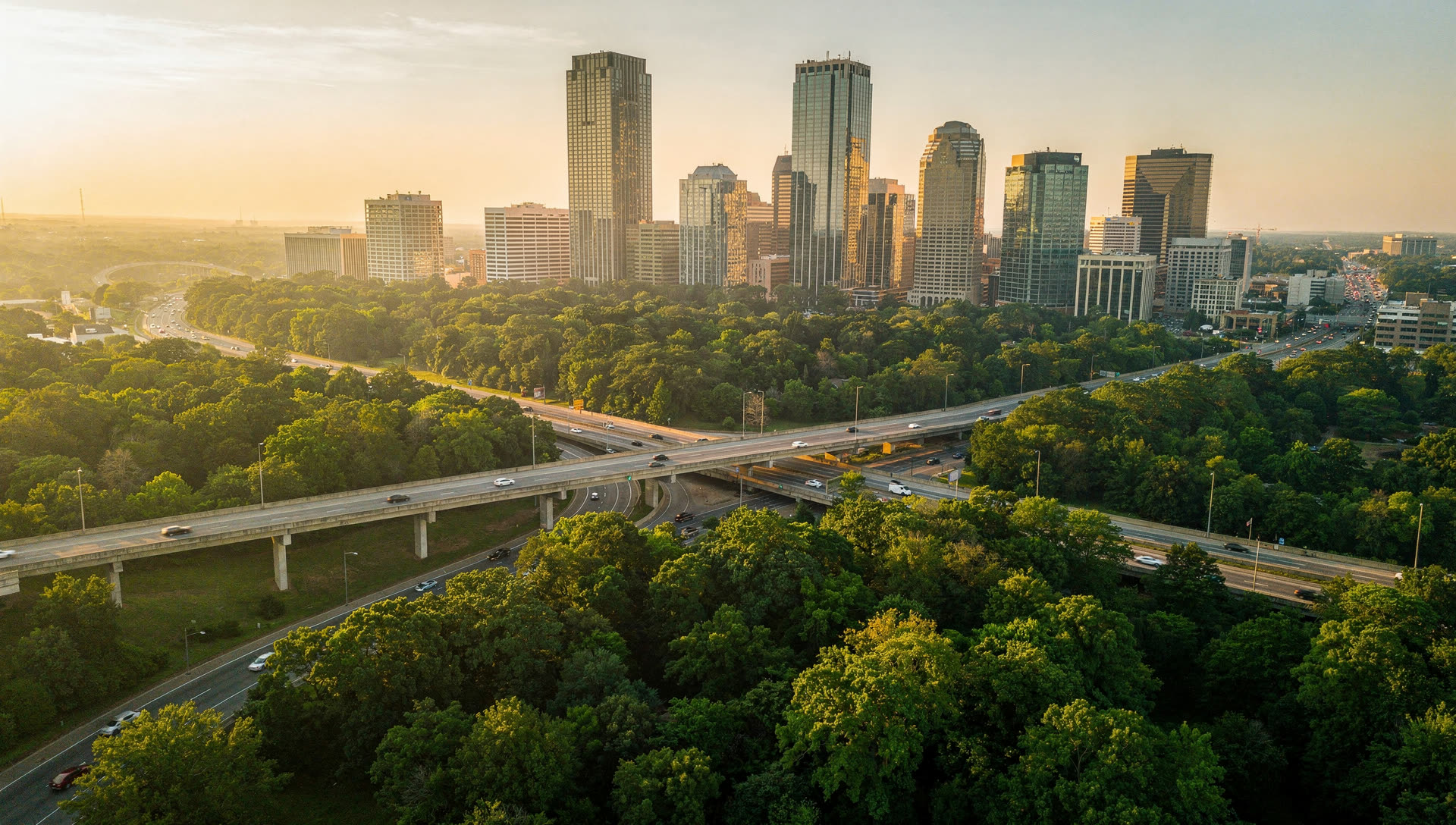 Aerial view over Atlanta at golden hour, Midtown and Downtown towers emerging from an unusually dense urban tree canopy with an interstate interchange threading between them.