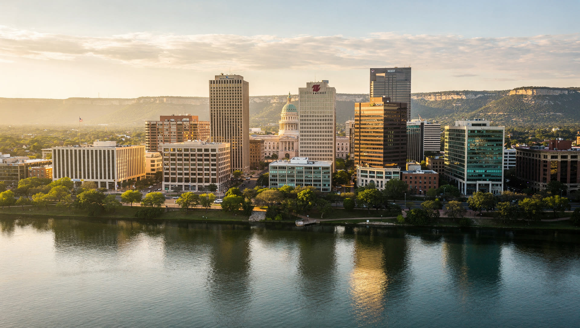 Austin, Texas from the air at golden hour, the downtown towers along Lady Bird Lake with the Colorado River curving east and the Hill Country on the western horizon.