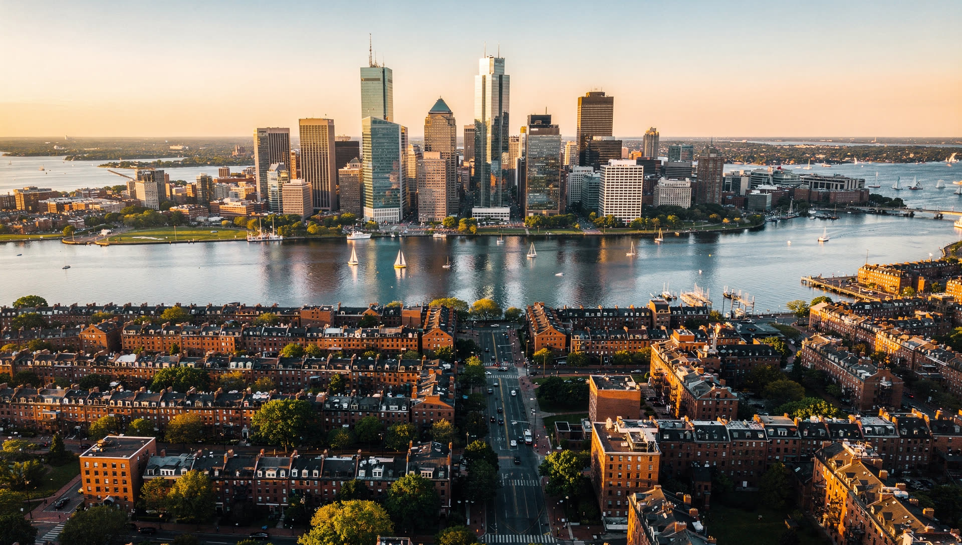 Boston from the air at golden hour, the Back Bay and Financial District towers along the Charles River with the harbor beyond.