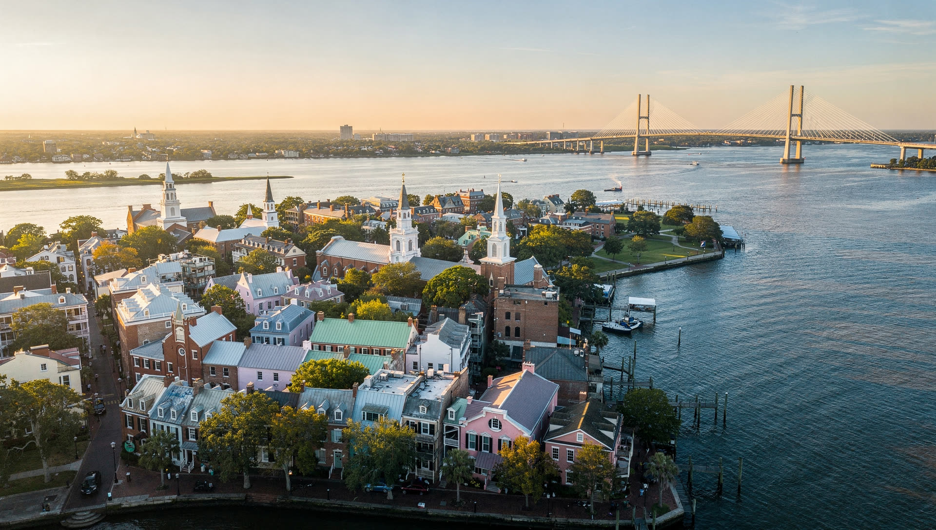 Aerial view over the Charleston peninsula at golden hour, church steeples and pastel historic rooftops between the Ashley and Cooper rivers with the Ravenel Bridge cables beyond.