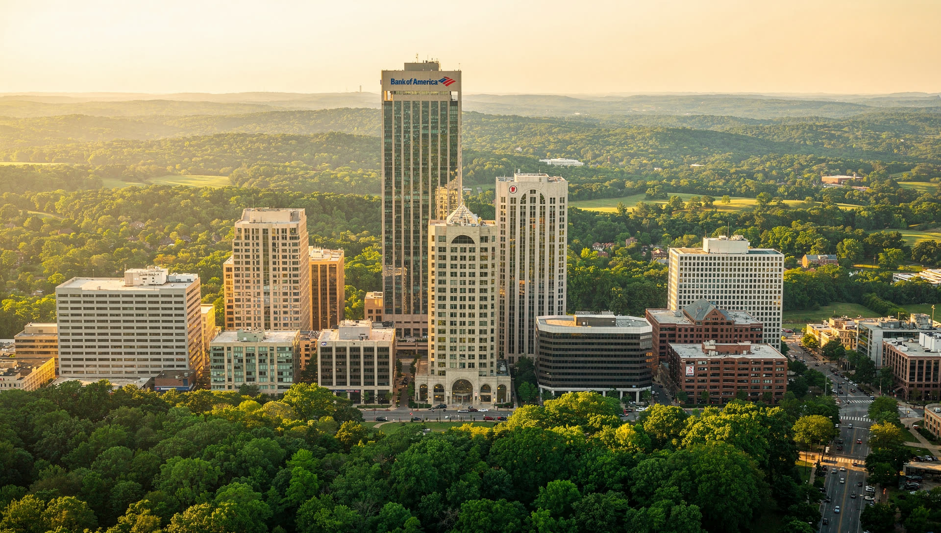 Aerial view over uptown Charlotte at golden hour, the banking towers rising from a dense tree canopy with the low rolling Piedmont beyond.