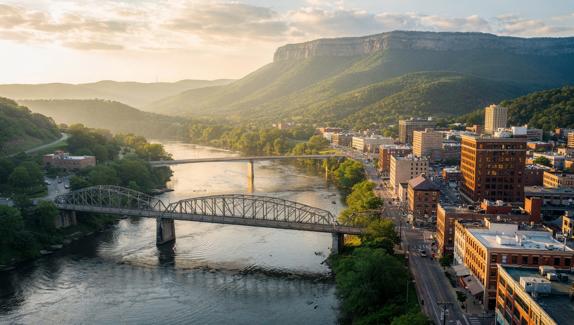 Aerial view over Chattanooga at golden hour, the downtown skyline in the Tennessee River gorge with the Walnut Street Bridge crossing and Lookout Mountain rising behind.