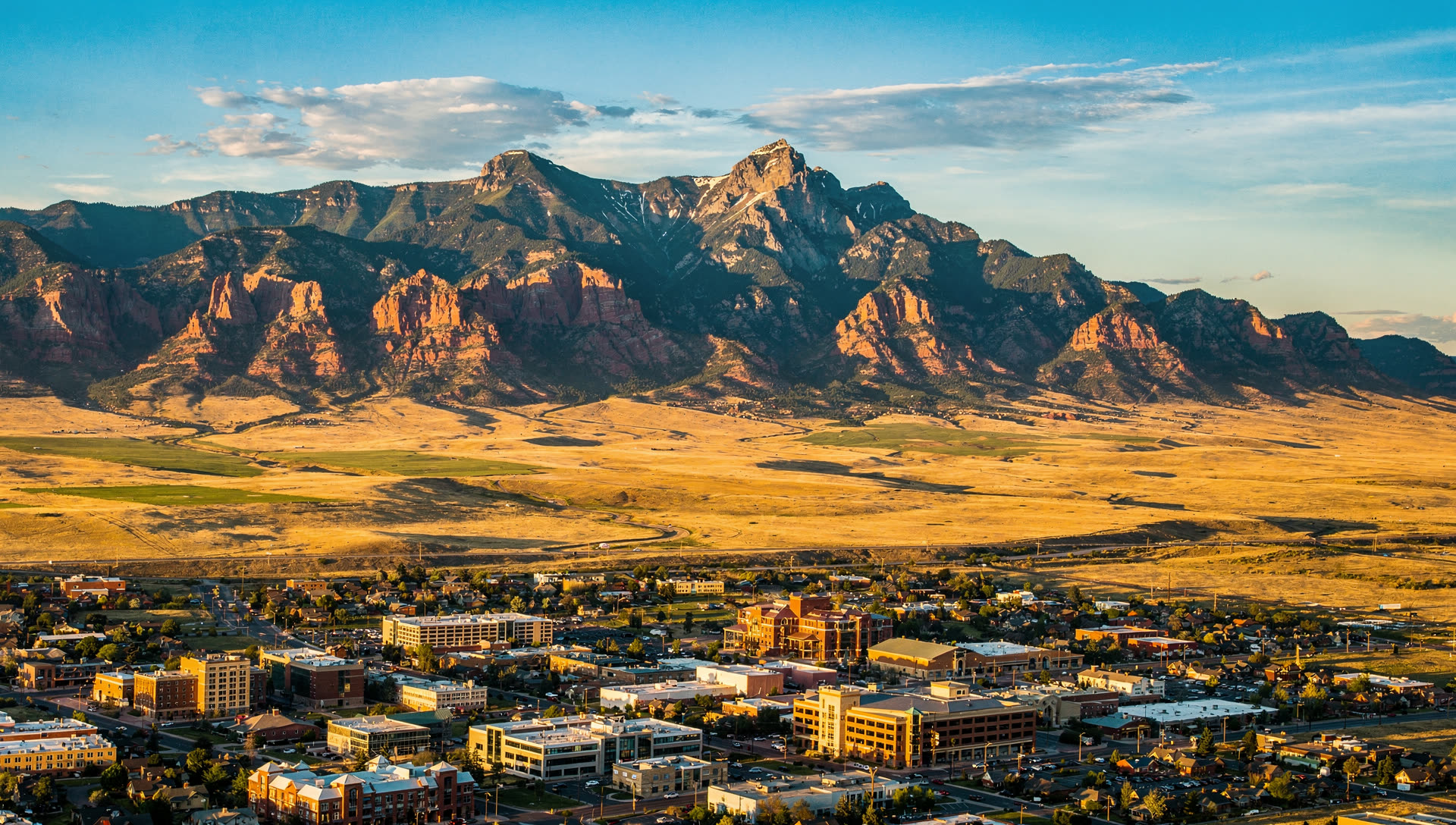 Aerial view of Colorado Springs at golden hour, the low downtown skyline on the plain with Pikes Peak and the Front Range rising steeply to the west.