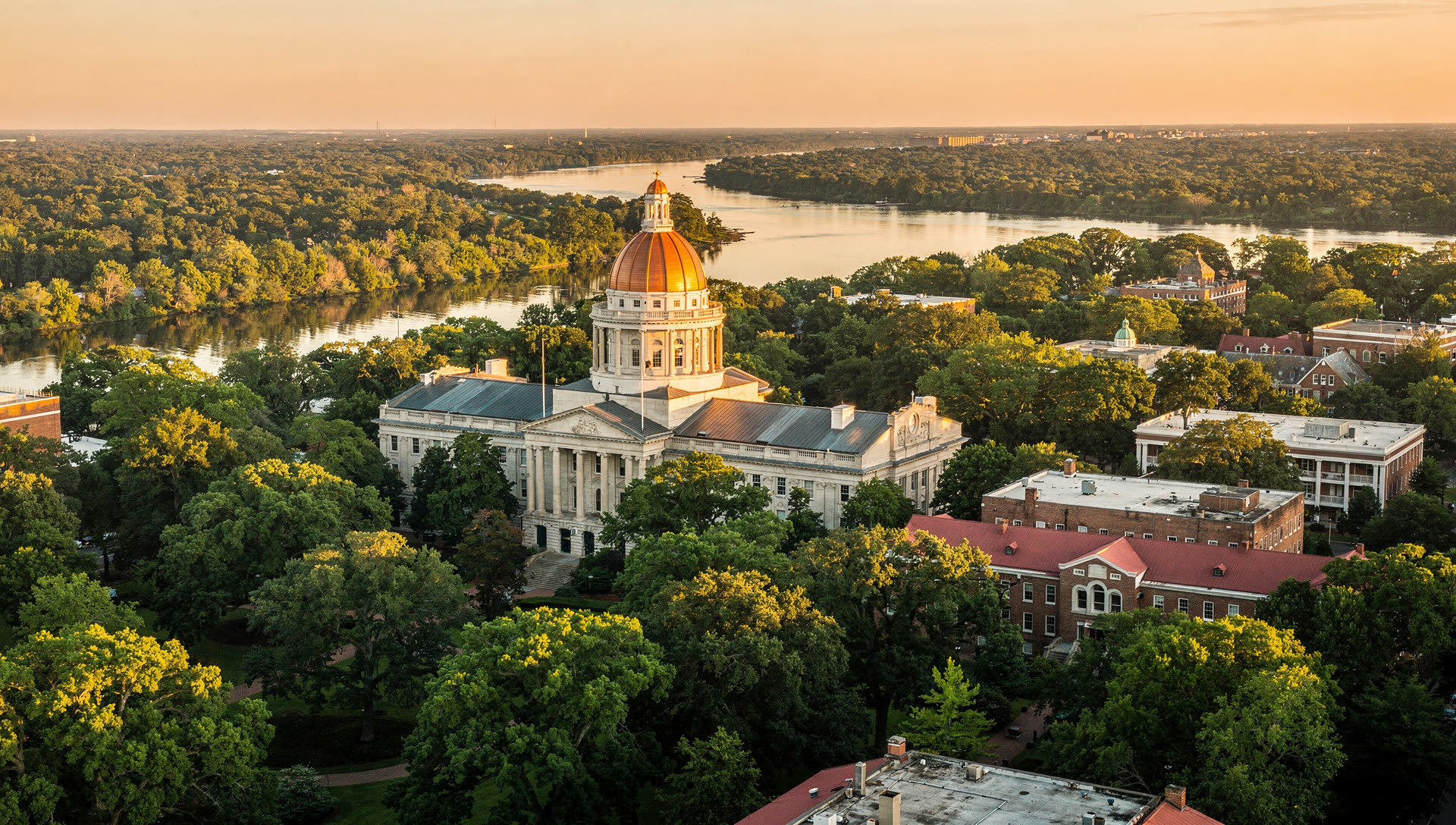 Aerial view over Columbia, South Carolina at golden hour, the State House dome and university campus above the Congaree River with dense southern hardwood canopy beyond.