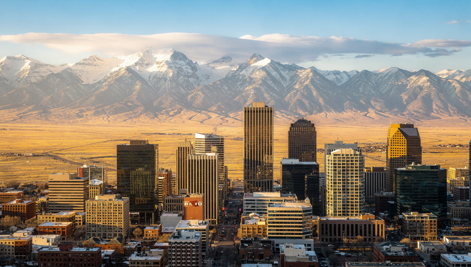 Aerial view of the Denver skyline at golden hour, downtown towers on the high plains with the Front Range of the Rocky Mountains rising along the western horizon.