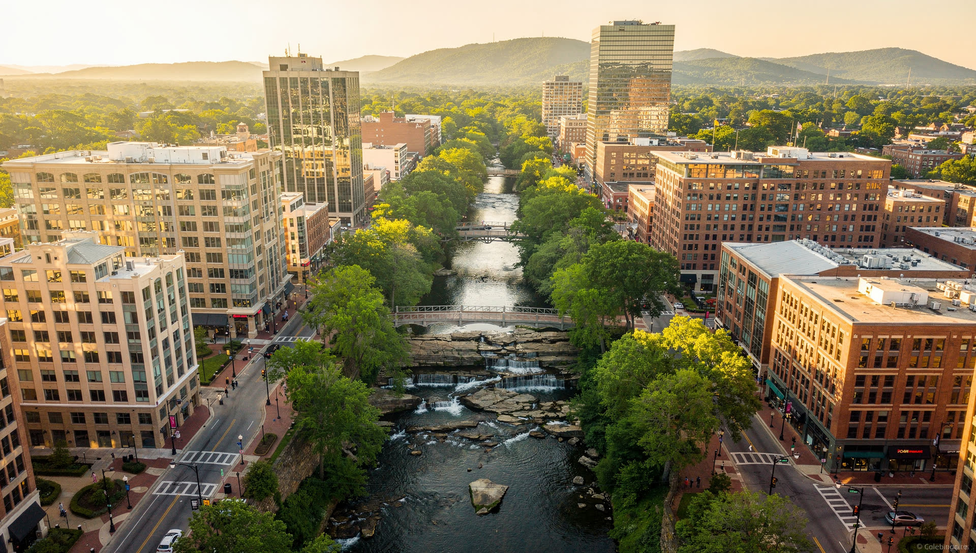 Aerial view over downtown Greenville, South Carolina at golden hour, the Reedy River falls and Main Street tree canopy in the foreground with the Blue Ridge foothills on the horizon.