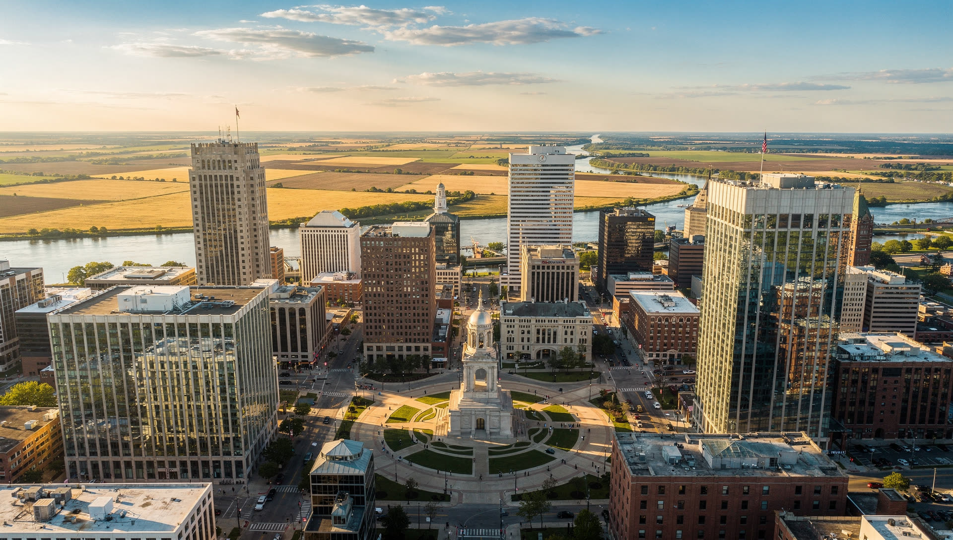 Aerial view of Indianapolis at golden hour, the downtown skyline and Monument Circle at the center of a flat grid of streets running to the horizon.