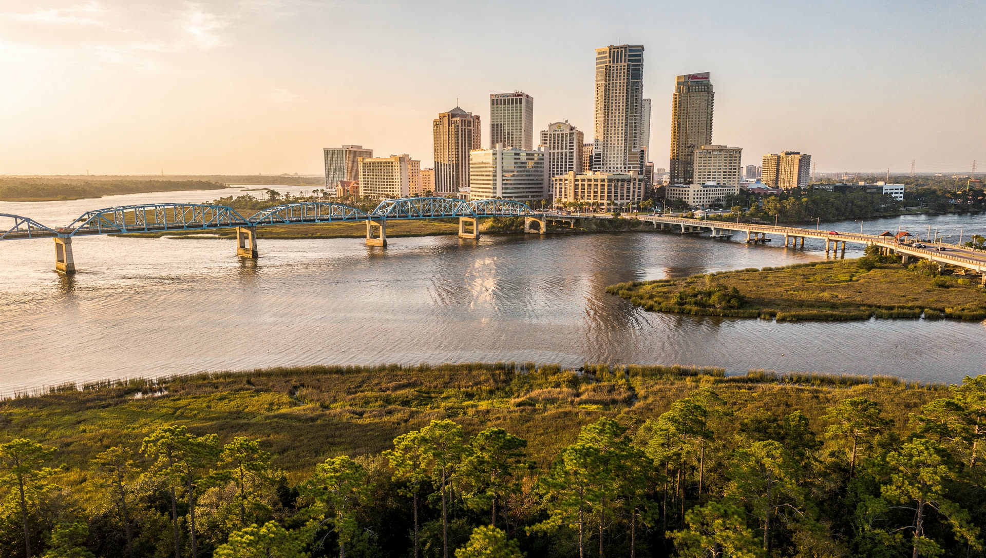 Jacksonville from the air at golden hour, the downtown towers on the St. Johns River with the bridges crossing to the southbank and the Atlantic beyond.