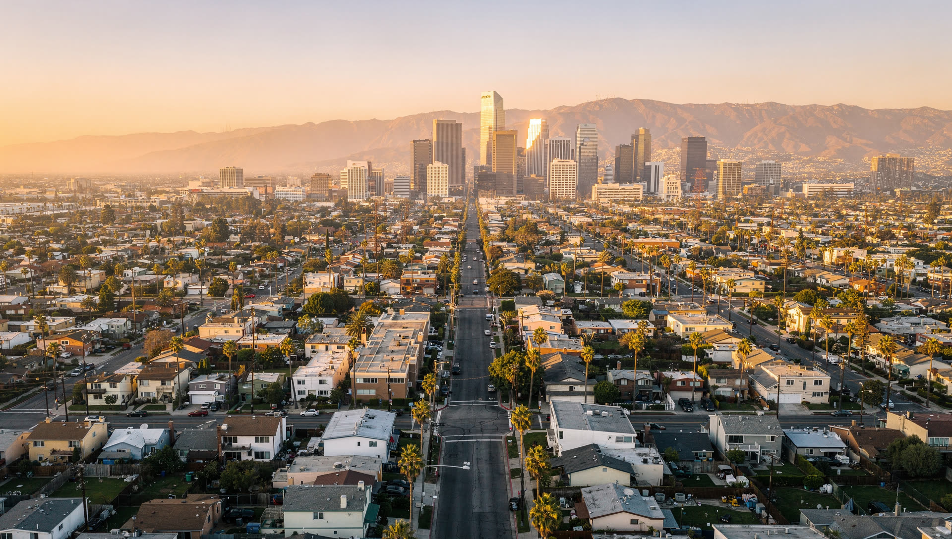 Aerial view over the Los Angeles basin at golden hour, downtown towers in the middle distance, the San Gabriel Mountains on the horizon, and a dense low-rise residential grid of palm-lined streets in the foreground.