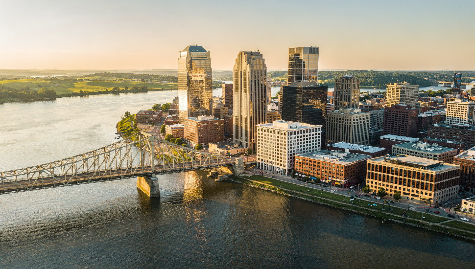 Aerial view of Louisville at golden hour, the downtown skyline on the Ohio River with the bridges to Indiana and the street grid running south and east.