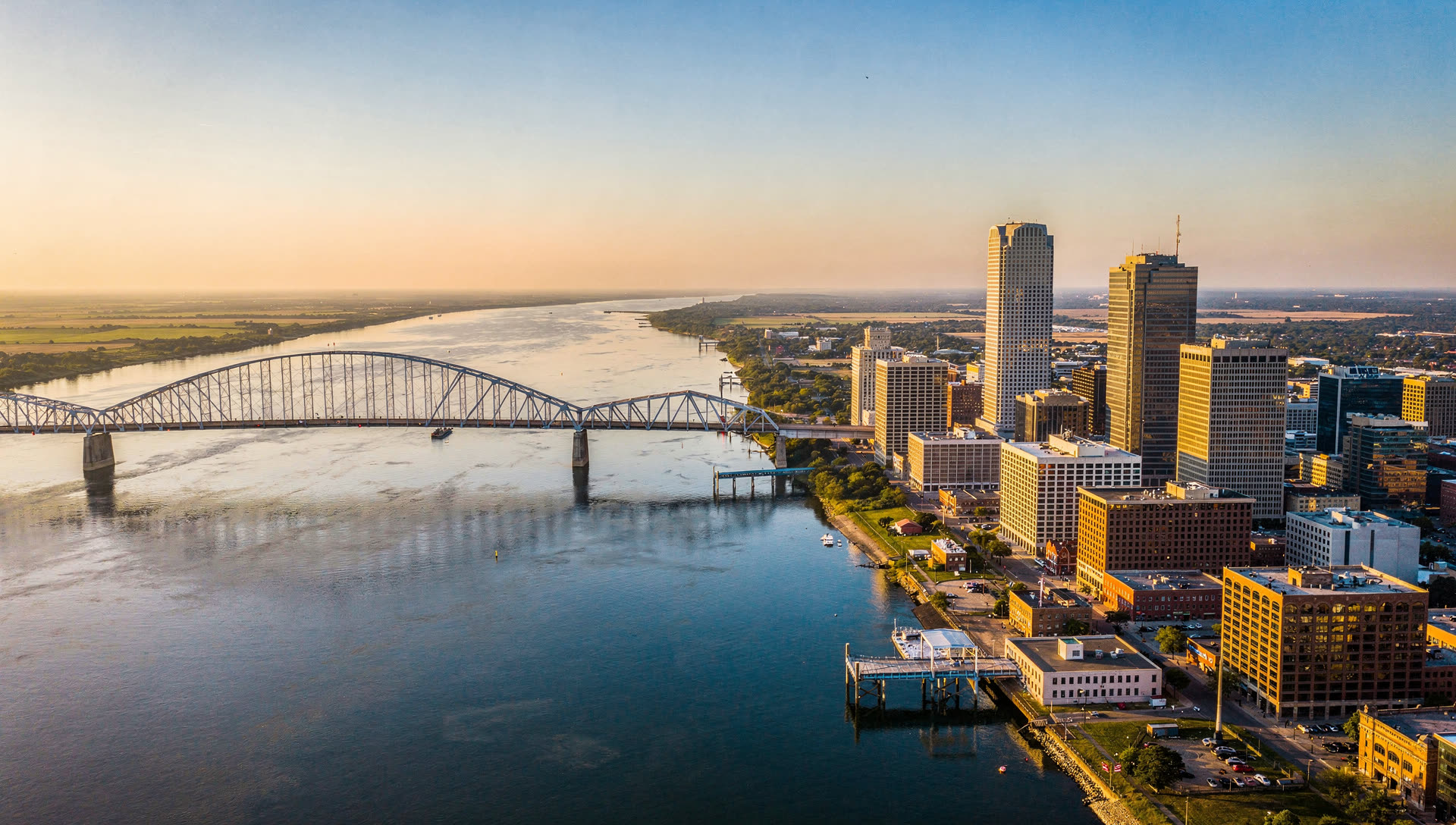 Memphis from the air at golden hour, the downtown towers on the Mississippi River bluff with the Hernando de Soto Bridge crossing into Arkansas.