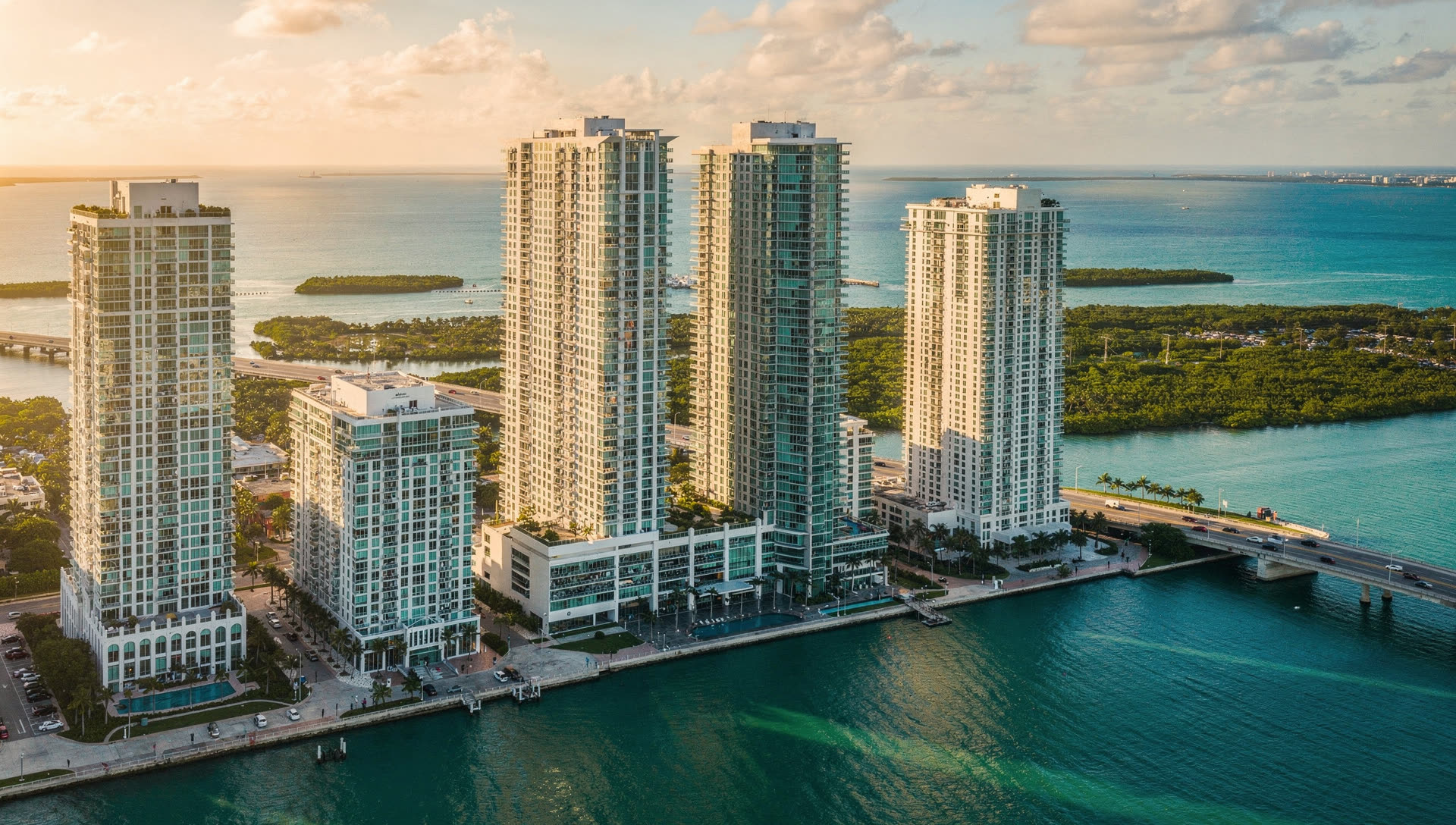 Aerial view over Miami at golden hour, the Brickell towers along Biscayne Bay with causeways crossing to the barrier islands and the Atlantic beyond.