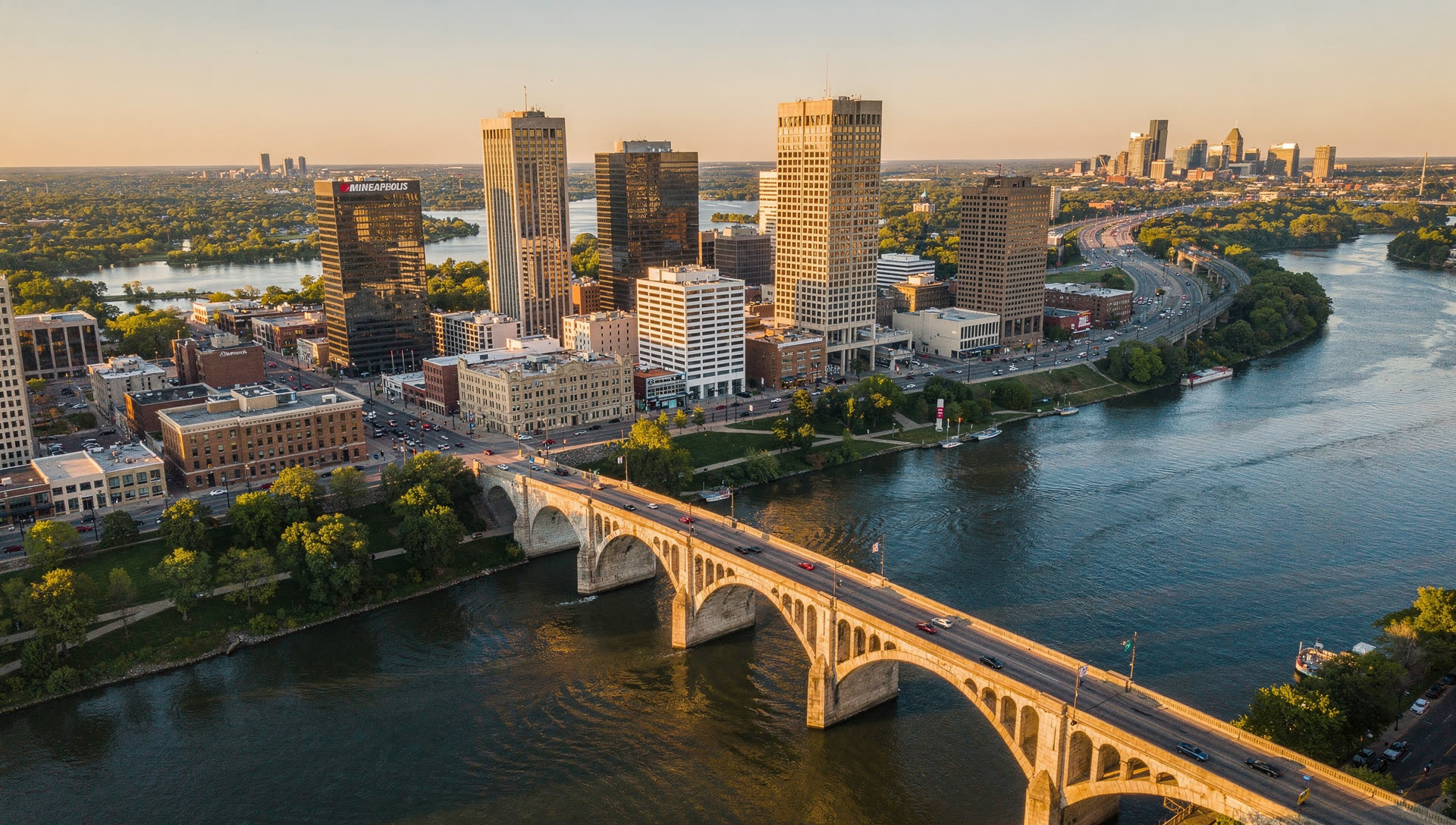 Aerial view of the Minneapolis and St. Paul skylines at golden hour, the Mississippi River curving between the two downtowns with the Stone Arch Bridge in the foreground.