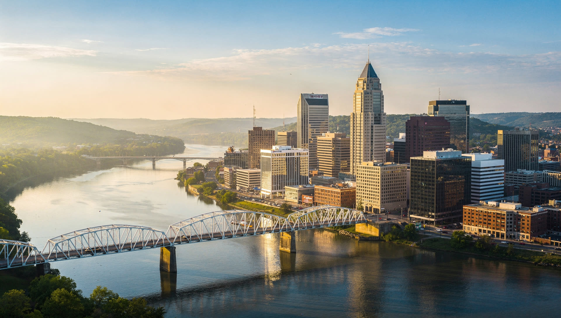Aerial view over Nashville at golden hour, the downtown skyline on the bend of the Cumberland River with a pedestrian truss bridge crossing and rolling Tennessee hills behind.