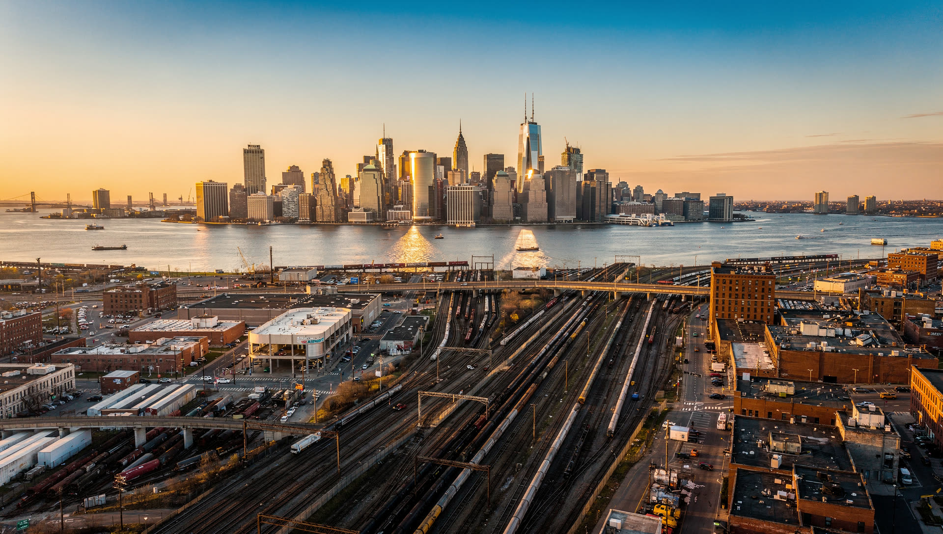Northern New Jersey from the air at golden hour, the Jersey City and Hoboken waterfront along the Hudson with the Meadowlands and the Watchung ridge beyond.