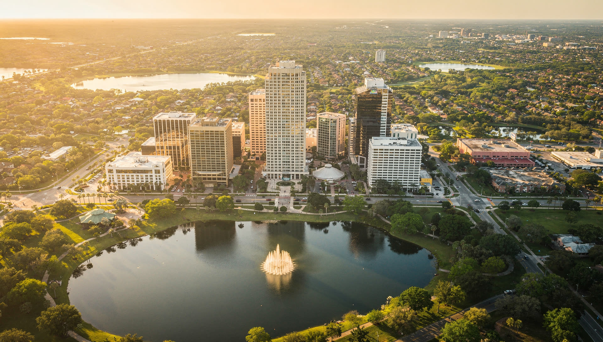 Orlando from the air at golden hour, the downtown towers beside Lake Eola with the chain of lakes and flat central Florida landscape stretching to the horizon.