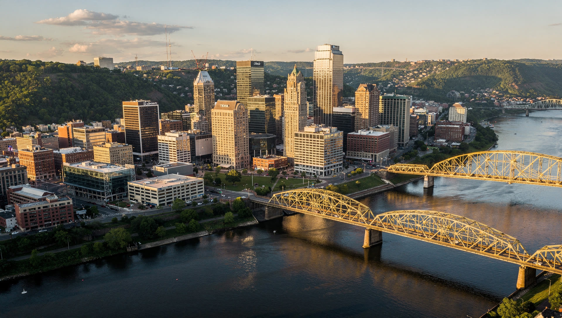 Aerial view of Pittsburgh at golden hour, the downtown triangle where the Allegheny and Monongahela rivers meet to form the Ohio, with bridges and hillsides around it.