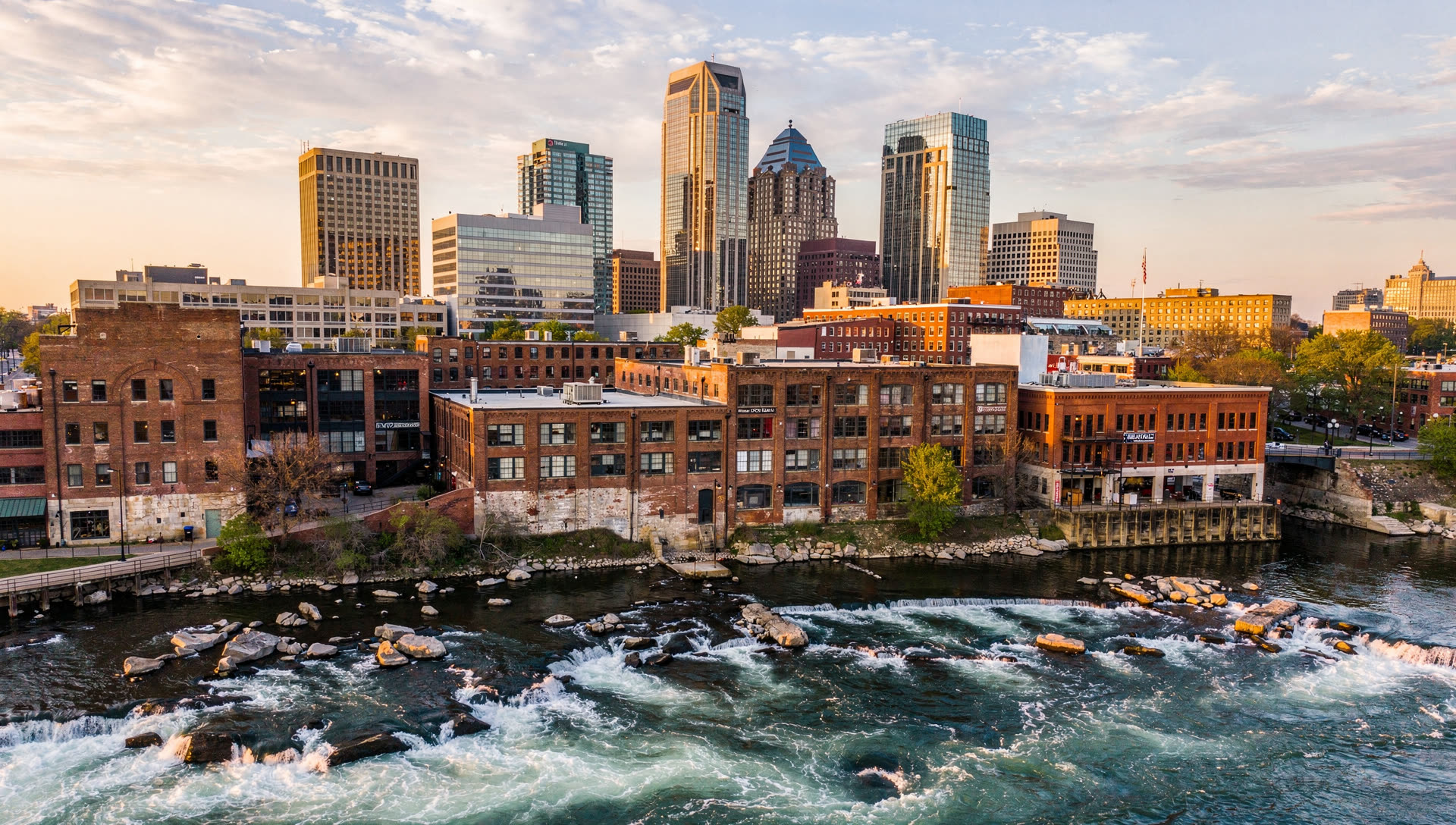 Aerial view over Richmond, Virginia at golden hour, the downtown skyline above the James River rapids with the historic brick warehouse district in the foreground.