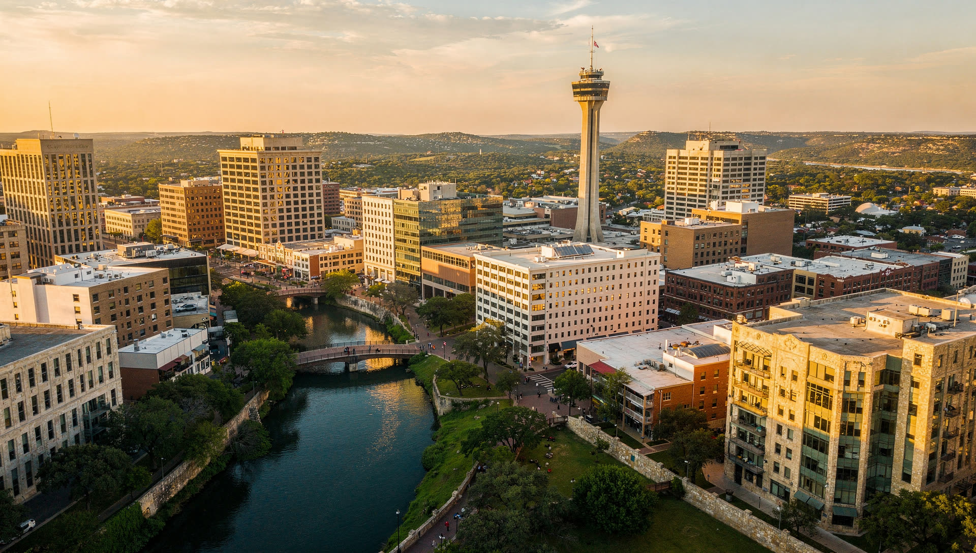 Aerial view of San Antonio at golden hour, the downtown skyline above the San Antonio River with the Tower of the Americas and low-rise neighborhoods spreading south and west.