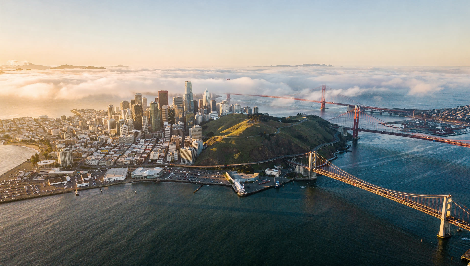 The San Francisco Bay Area from the air at golden hour, the downtown San Francisco towers with the Bay Bridge crossing to Oakland and the East Bay hills beyond.