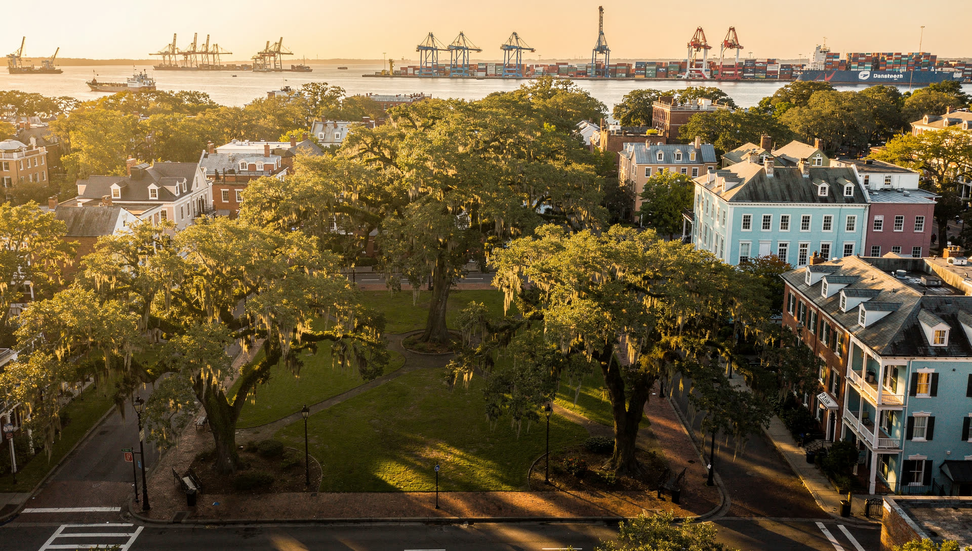 Aerial view over Savannah at golden hour, the historic district grid of live-oak squares draped in Spanish moss with the Savannah River and container terminal beyond.