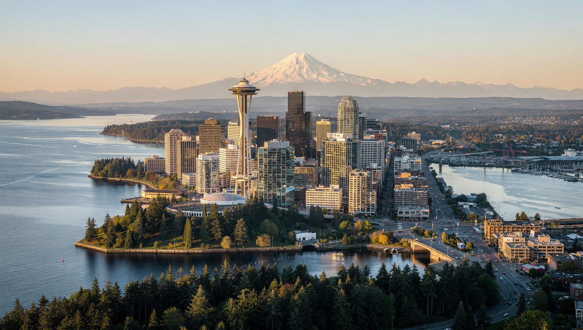 Aerial view of Seattle at golden hour, the downtown towers on Elliott Bay with Lake Union behind and Mount Rainier on the southern horizon.