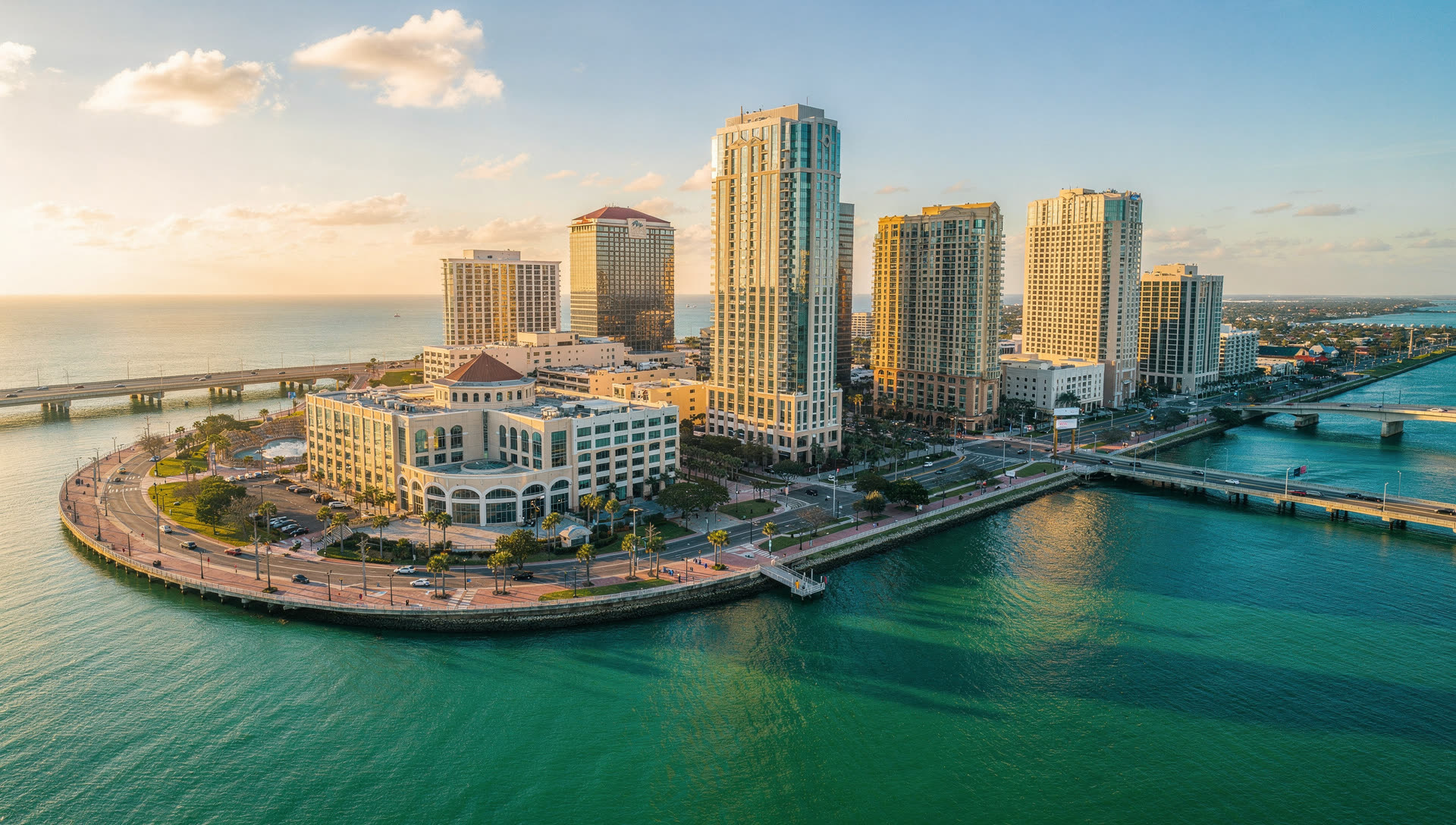Aerial view over Tampa at golden hour, the downtown skyline on Hillsborough Bay with the Riverwalk curve in the foreground and causeways crossing the water beyond.